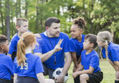 Six multi-ethnic children 6-7 years old playing soccer at summer camp. They are gathered around their coach who is talking strategy.